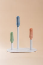 Three silicone finger brushes in green, blue, and orange, displayed on a white stand against a solid beige background.