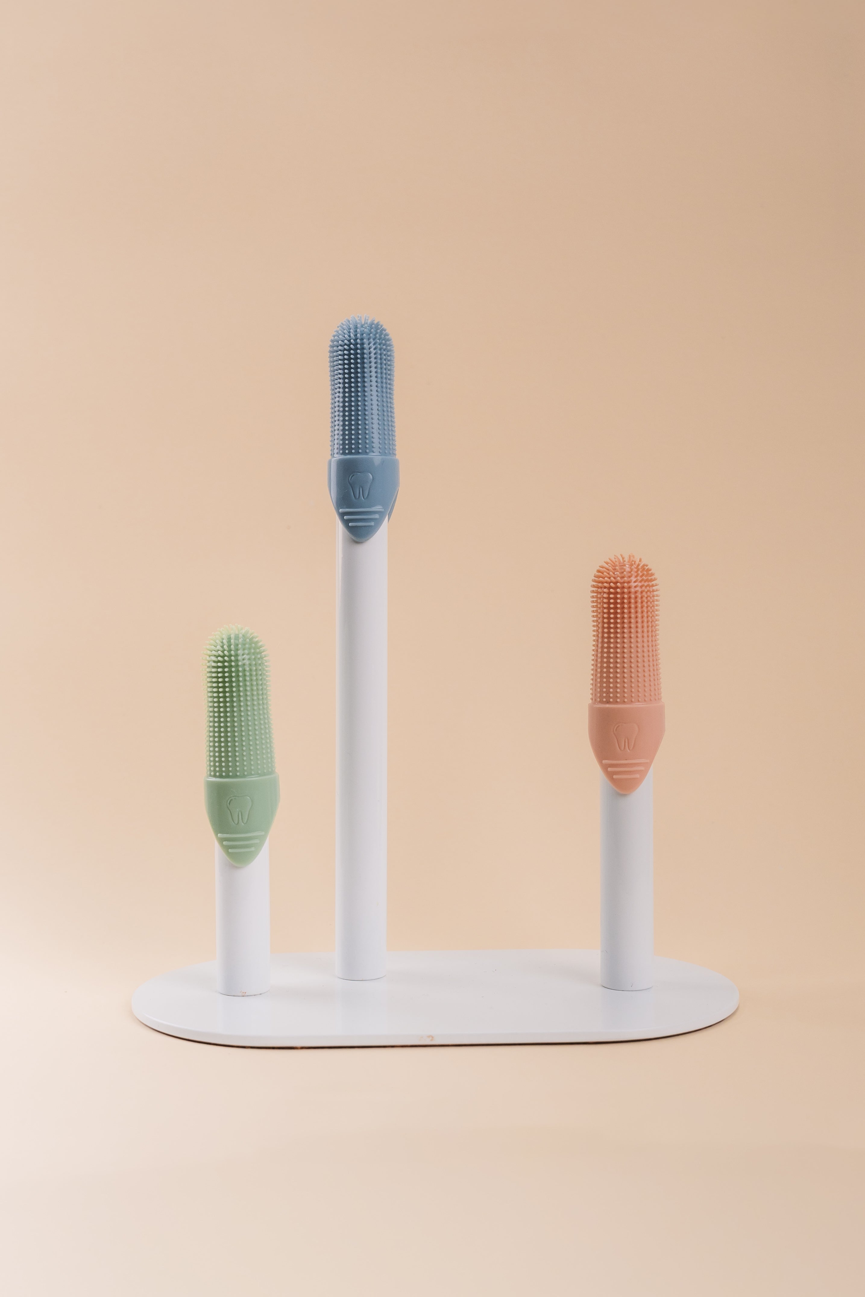 Three silicone finger brushes in green, blue, and orange, displayed on a white stand against a solid beige background.