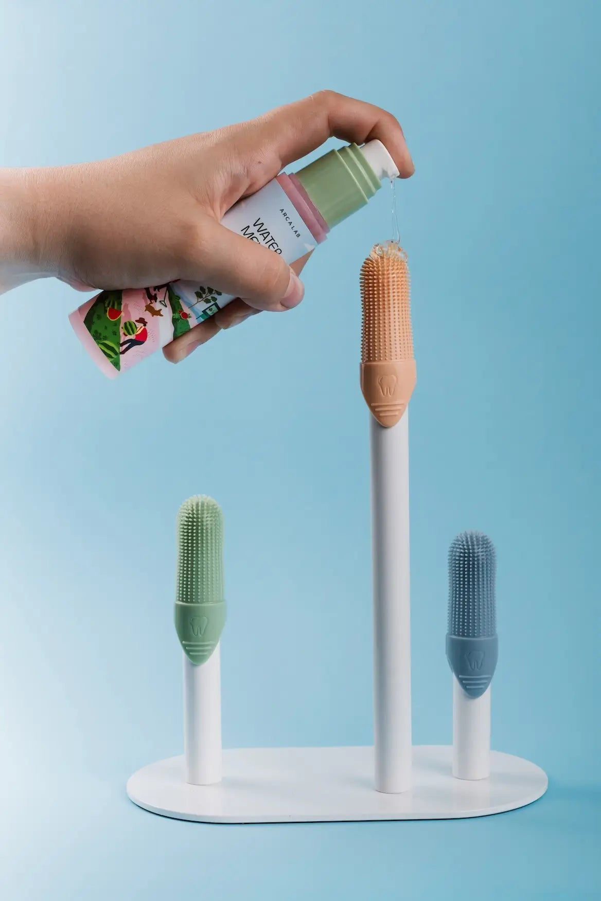 A hand holds a bottle of hand wash over a white stand holding three silicone cleaning finger brush heads in crisp green, orange, and blue, against a light blue background.