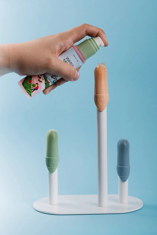 A hand holds a bottle of hand wash over a white stand holding three silicone cleaning finger brush heads in crisp green, orange, and blue, against a light blue background.