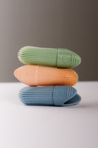 Three silicone oral care finger brushes stacked vertically in crisp green, orange, and sky blue, featuring textured bristles and a tooth icon embossed on the handle.