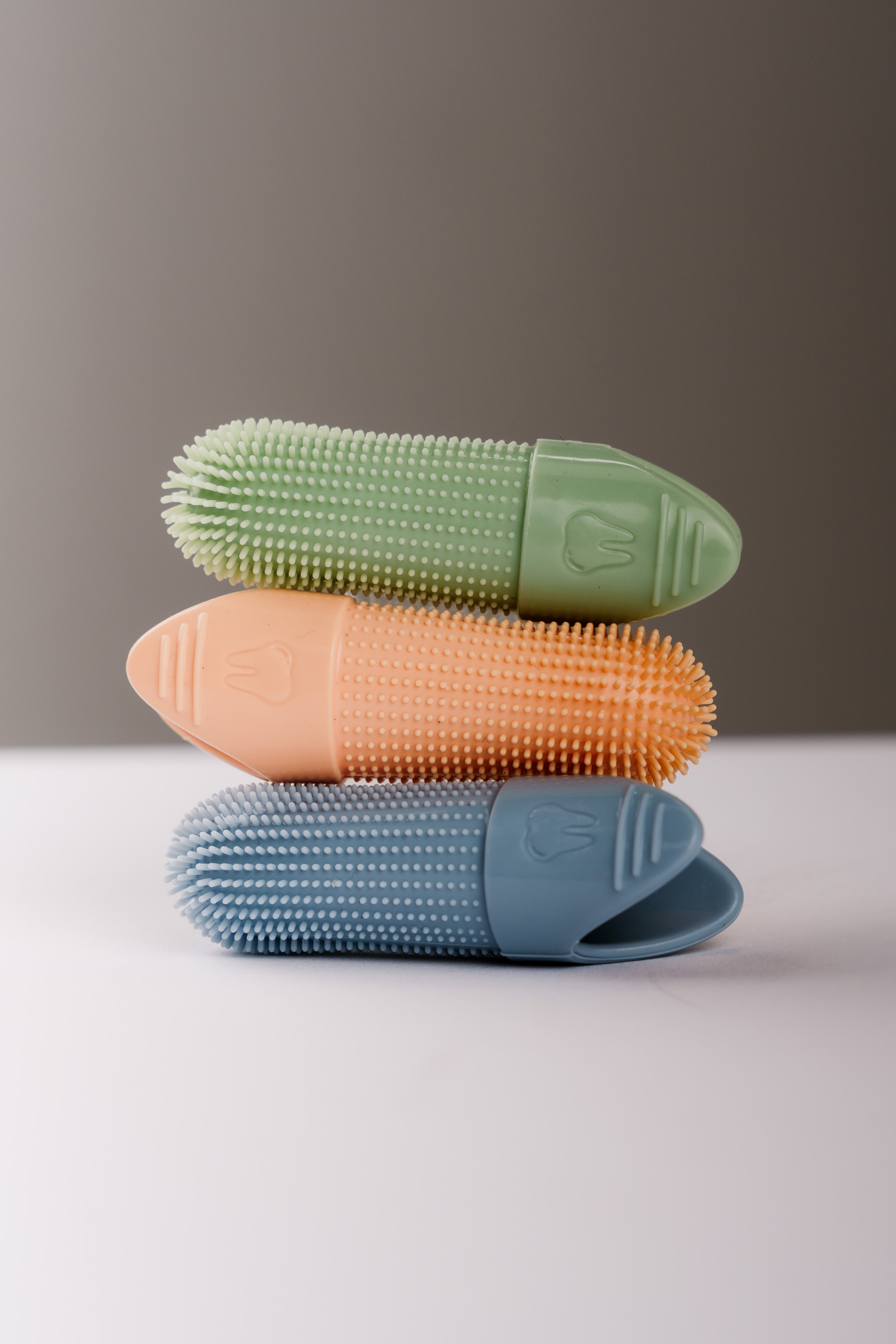 Three silicone oral care finger brushes stacked vertically in crisp green, orange, and sky blue, featuring textured bristles and a tooth icon embossed on the handle.