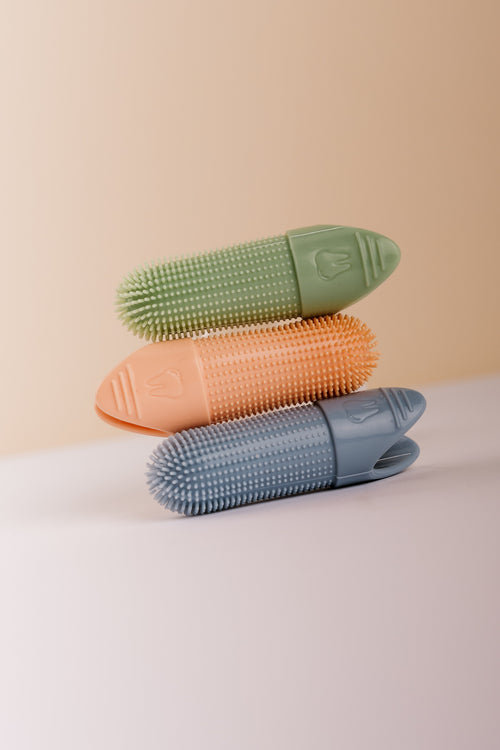 Three textured silicone finger brush accessories stacked vertically in green, orange, and blue on a clean, two-toned beige and white surface.