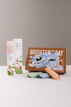 A dog toothpaste gift set from Arca Lab on a light gray surface against a neutral gray background. The set includes a Watermelon Burst Enzymatic Toothpaste bottle, its box, a brown illustrated cardboard gift mailing box reading 'BRIGHTEN YOUR DAY', and three finger brushes in crisp green, blue, and orange.